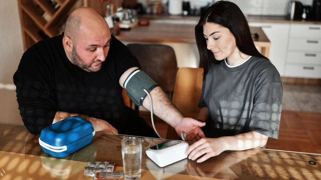 A woman takes the blood pressure of a man with a blood pressure cuff at a table inside a house